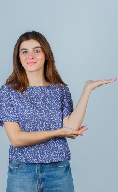 young girl spreading palm aside, holding hand under elbow in blue patterned t shirt, jeans and looking happy , front view.